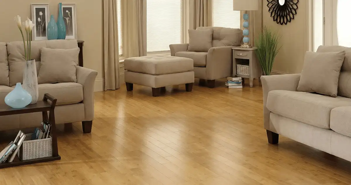 A living room with professionally installed bamboo flooring, beige sofas, a matching ottoman, a side table with a blue lamp, vases, framed pictures, and large windows with blinds and curtains in Los Angeles, CA