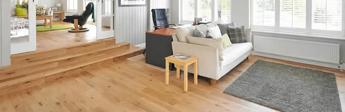 A modern living room with professionally installed laminate flooring, a beige sofa, a small side table, a gray rug, and large windows with white shutters in Los Angeles, CA
